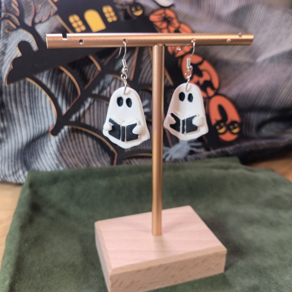 White ghost reading a black spell book dangle earrings on a gold display stand with Halloween backdrop