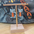 Grumpy grey and white Halloween cat dangle earrings with orange outfit on a gold display stand