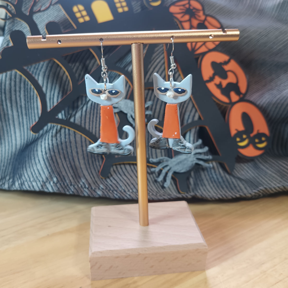 Grumpy grey and white Halloween cat dangle earrings with orange outfit on a gold display stand