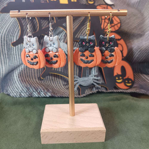 Grey and black cat in jack-o'-lantern dangle earrings displayed on a gold stand with Halloween backdrop