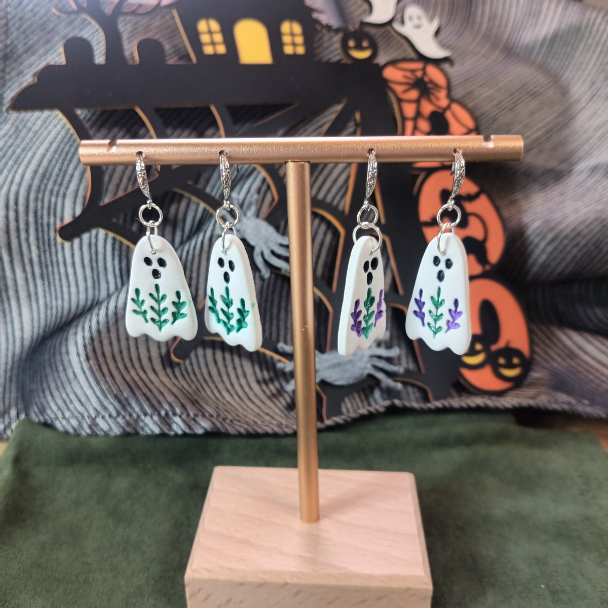 White ghost dangle earrings with green and purple botanical sprig details on a gold display stand with Halloween backdrop