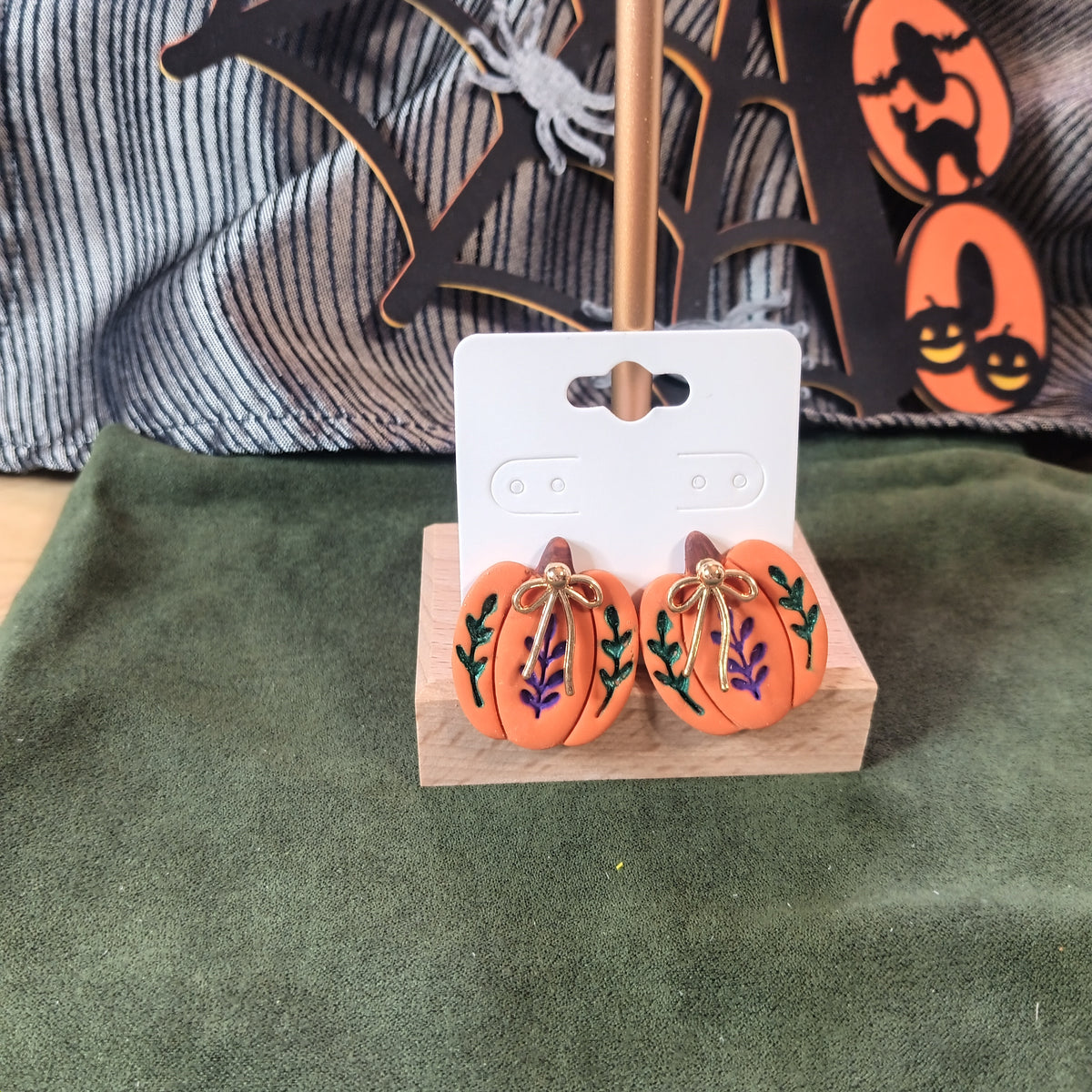 Orange pumpkin stud earrings with green and purple botanical details and gold bow charm on a wooden earring card stand