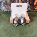 White ghost inside an ornate dark silver oval portrait frame dangle earrings on a wooden earring card stand