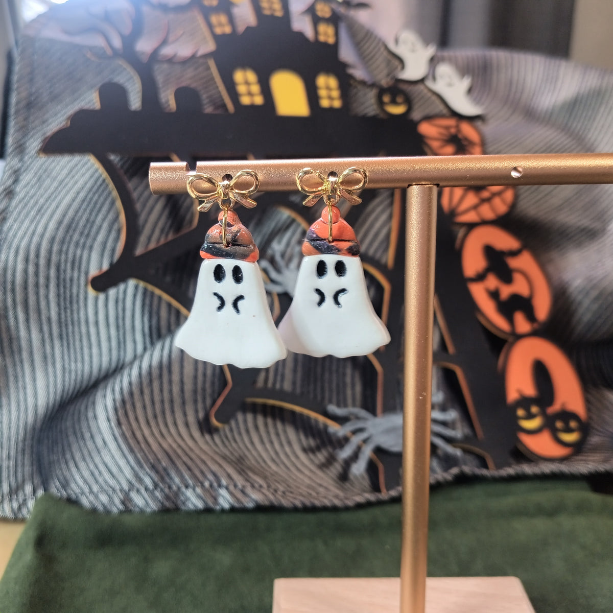 White ghost wearing orange and black witch hat dangle earrings with gold bow studs on a gold display stand