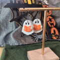 White ghost reading an orange book dangle earrings on a gold display stand with Halloween backdrop