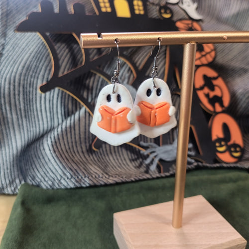 White ghost reading an orange book dangle earrings on a gold display stand with Halloween backdrop