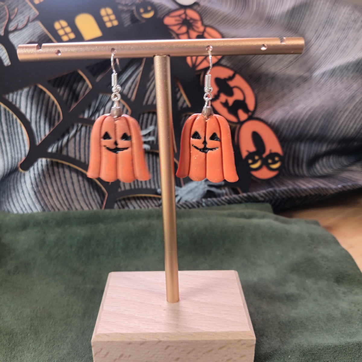 Orange pumpkin ghost dangle earrings hanging on a gold display stand with Halloween backdrop